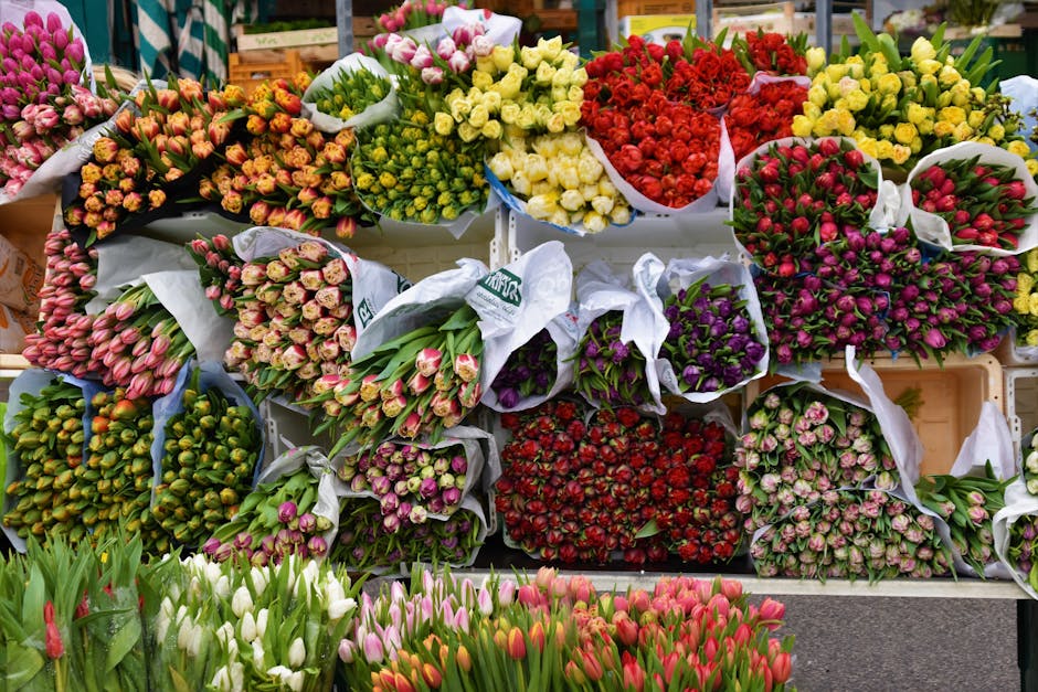 A vibrant display of freshly cut flowers arranged in bouquets and bundles at a flower market. The collection features tulips in shades of pink, white, and red at the front, with the tulips appearing c
