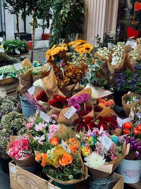 A vibrant display of fresh flower bouquets arranged outdoors, featuring a variety of blooms wrapped in brown paper and tied with white ribbons. The bouquets include bright yellow sunflowers, pink lili